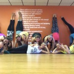 Group of women posing playfully with legs up against a wall in a dance studio.