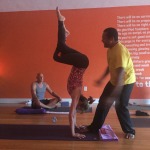 A man assists a woman in a yoga handstand with another person meditating nearby.
