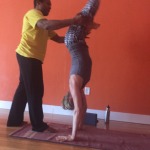 A person assists another in a handstand against an orange wall.