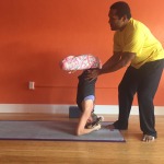 A man assists a child doing a headstand on a yoga mat indoors.