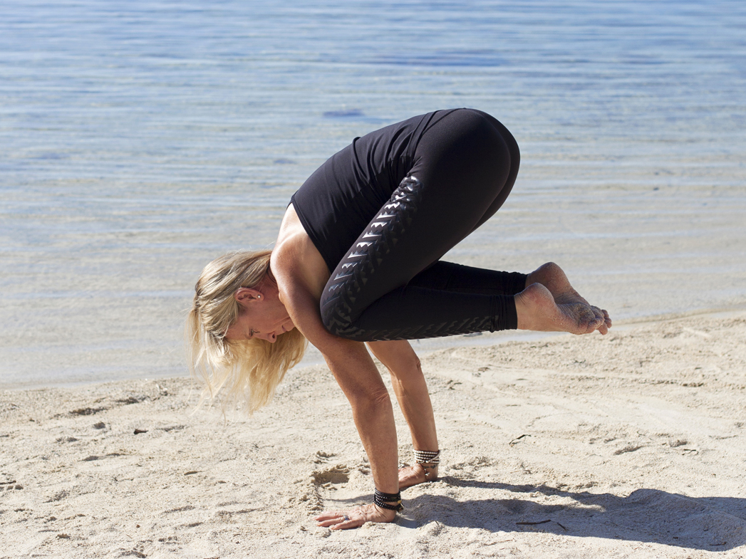 A woman performing a yoga crow pose on the beach near the water.